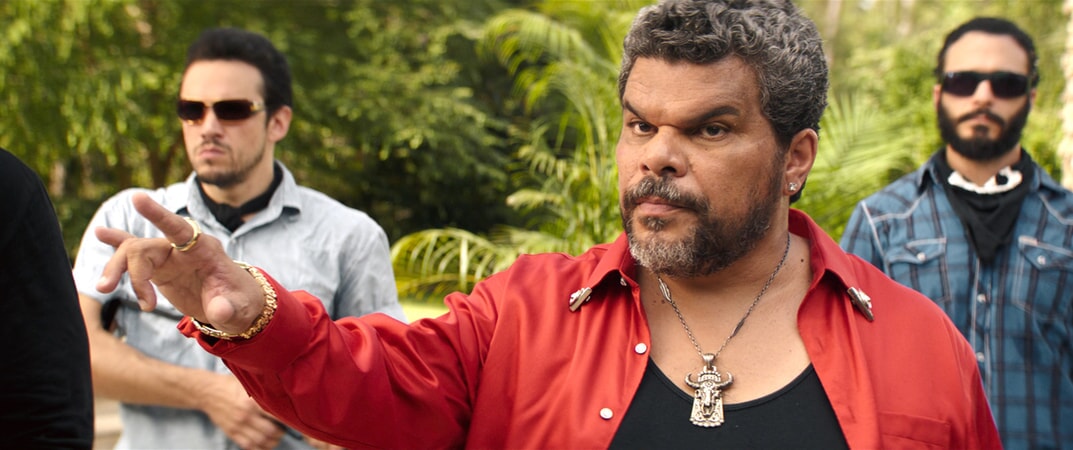 LUIS GUZMÁN as Bacon wearing a red jacket and amulet necklace
